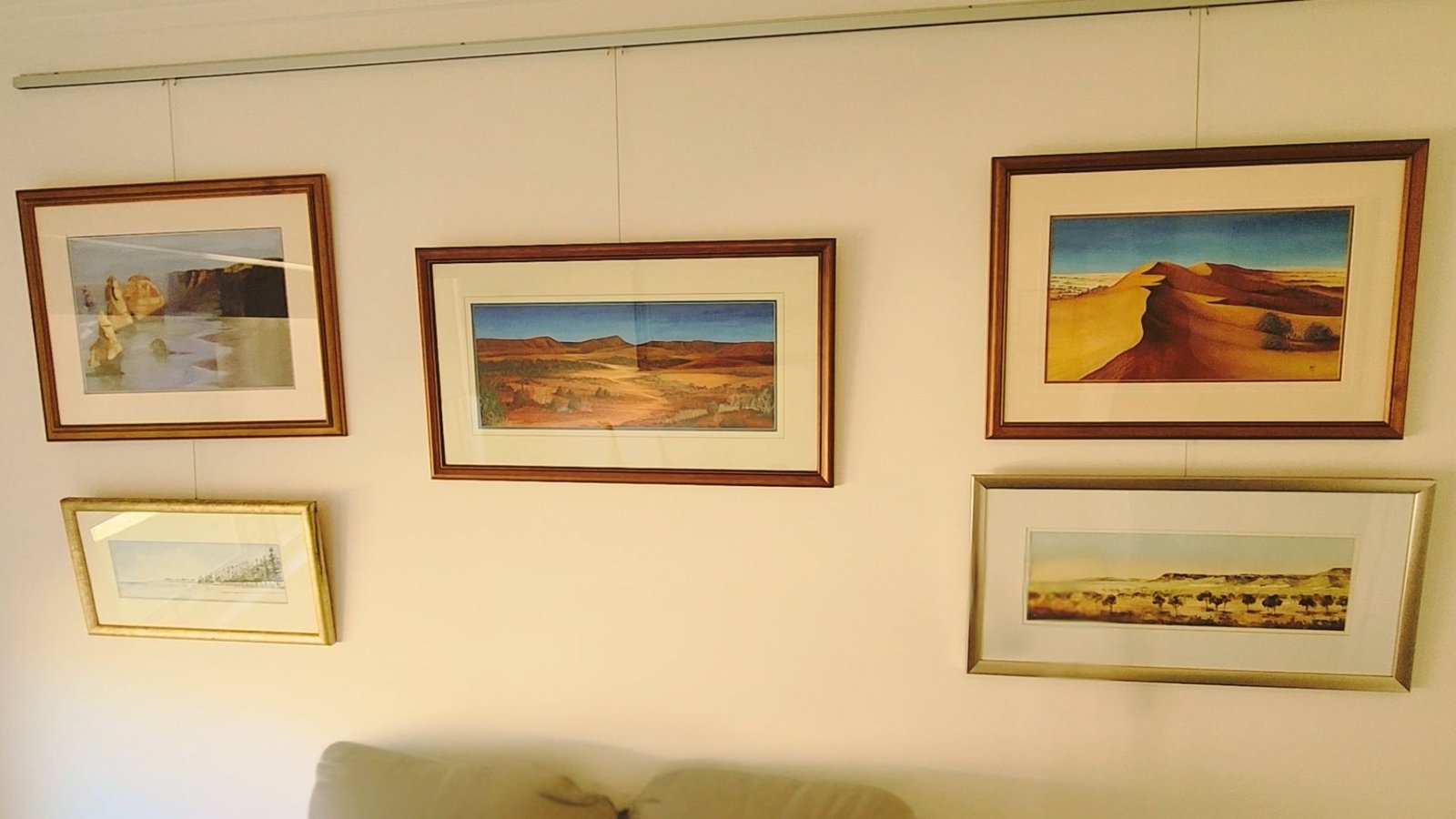 A wall displaying several framed paintings hung using a wire gallery system, showing the professional result of Cinnamon's art hanging service in customer Geoff's home.
