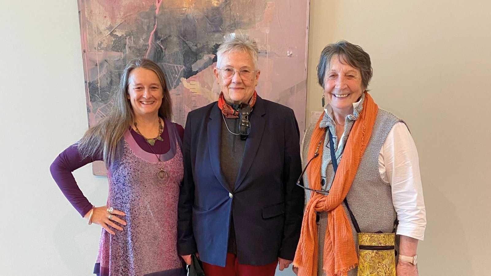 Cinnamon is smiling and standing between two other women in an art gallery space, visually representing her work supporting and connecting local artists and art lovers on the Surf Coast.