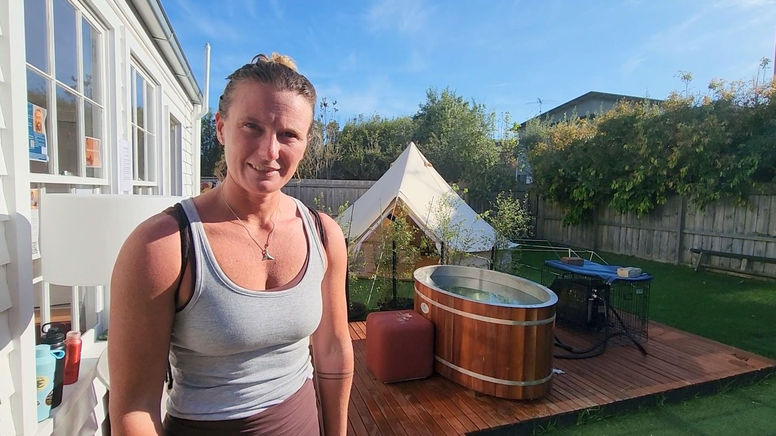 Club member Carly stands smiling on a sunny outdoor wooden deck next to a round ice bath and a white canvas tent at Sublime Health Club in Torquay.