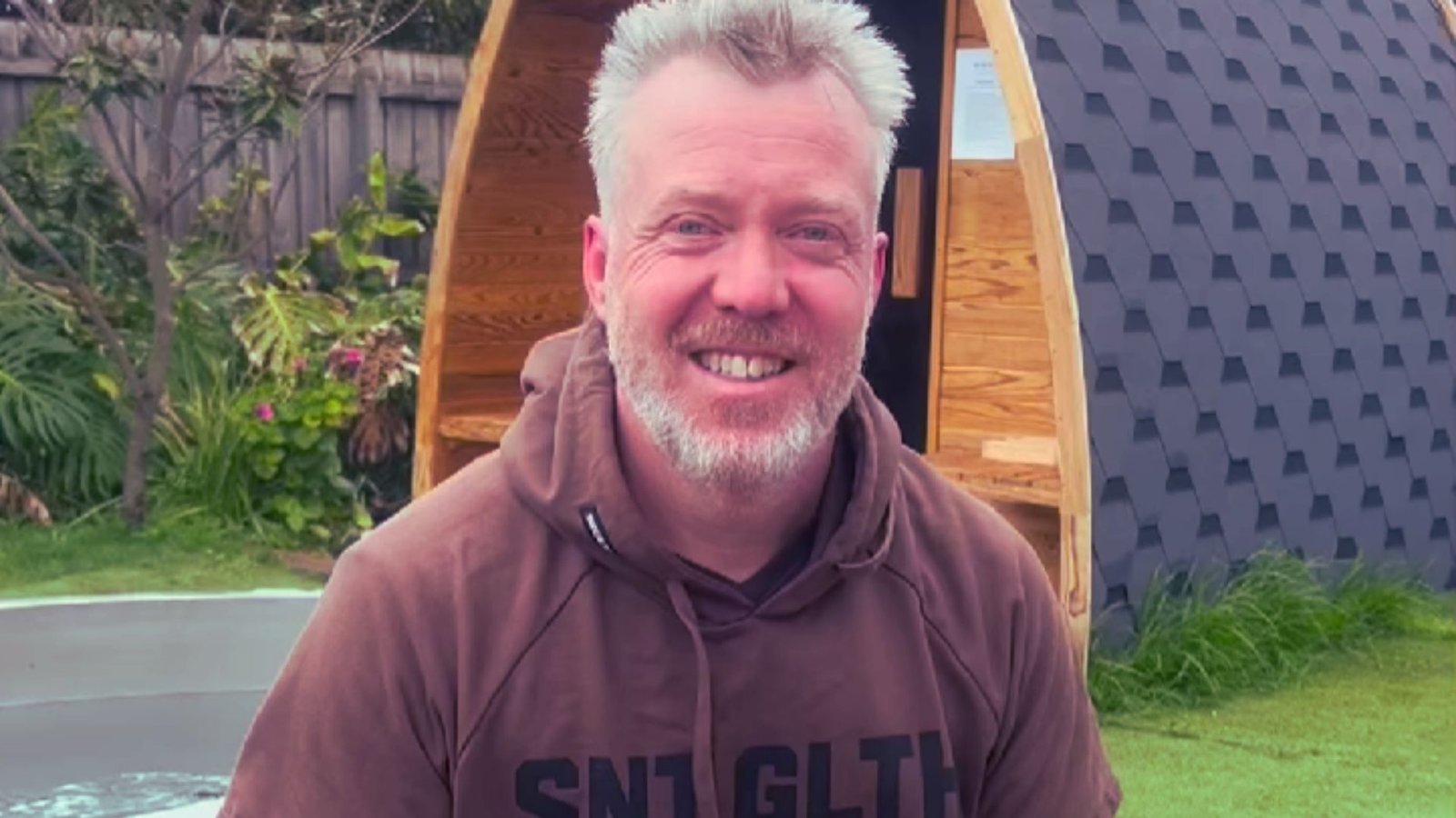 Club member Matt smiling warmly while wearing a brown hoodie, sitting outdoors on the green grass with a wooden barrel sauna visible in the background at Sublime Health Club in Torquay.